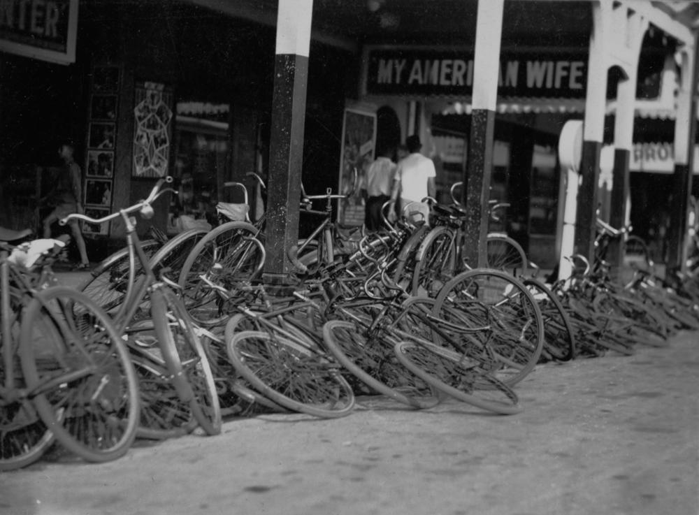 Black and white image showing numerous bikes that have been left on the street outside a cinema. Two people walk on the footpath outside the cinema, under a sign above which reads 'MY AMERICAN WIFE'