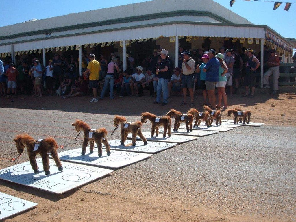 Colour photograph showing a row of nine small, toy horses on cardboard bases, lined up outside a hotel. A crowd of people has gathered in the background.
