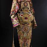 A long-sleeved embroidered cream silk jacket. The sleeves are in polished cotton with appliqued floral emblems and gold thread and silk embroidery.There are decorative panels attached over the shoulders and ties at waist level to attach the front and back sections. Below this the front and back panels are not attached to each other. In the upper front section there is a dragon face with a raised purple nose and red ears trimed with white fur. The lower front and back panels are lined with red cloth while the upper section is lined with cream cloth. The lower front panel has a double decorative layer.
