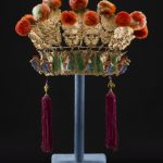 An elaborate Chinese headdress made up of a lower section painted in gold, red, green and blue. Above this there is a row of gold floral emblems to which are attached red pompoms.