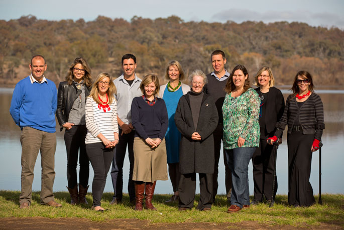 Eleven people stand in two rows on grass, in front of a watercourse, and with trees in the background.