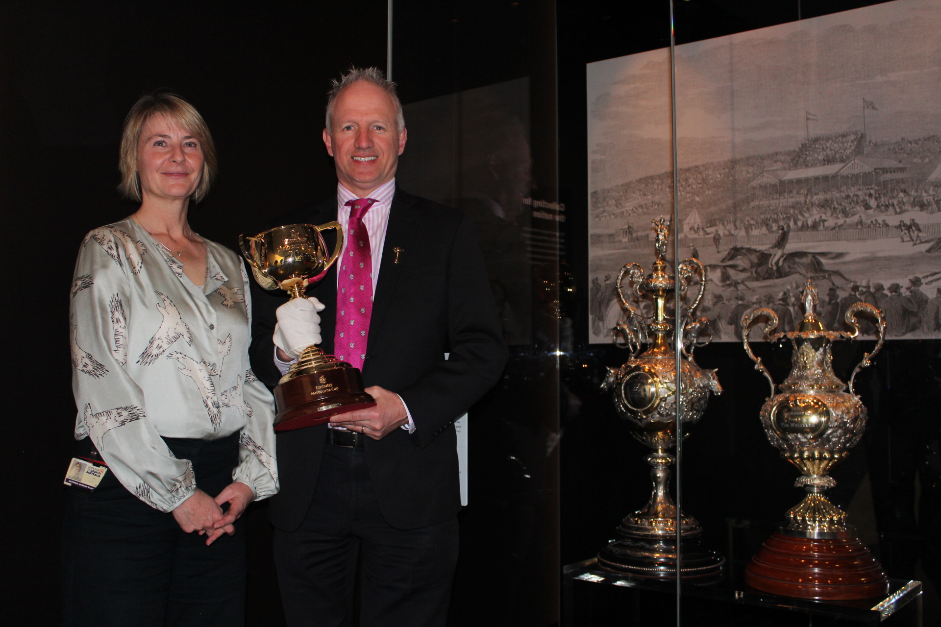 A man holding a gold cup and a woman stand next to a display case holding two ornate silver trophies.
