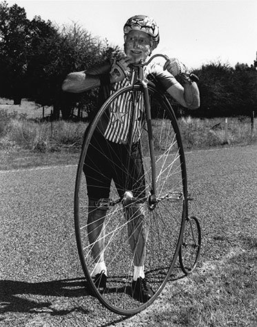 Black and white photograph of a man standing beside a penny-farthing bicycle. The man, who wears a helmet and has zinc cream across his cheeks, rests his head on the handlebars. He wears cycling gloves, shorts and a shirt with stars and stripes at the front.