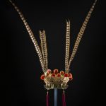 An elaborate Chinese headdress made up of a lower section painted in gold, red, green and blue. Above this there is a row of gold floral emblems to which are attached red pompoms and long speckled feathers.