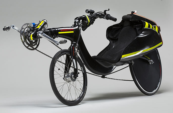 A recumbent bicycle with carbon fibre tailbox and two different size wheels. The rear wheel is larger and and has a black wheel cover. The pedals are in front of the front wheel. The steering is above seat. The body of the bicycle is black in colour with some bright yellow stripes. The text ' Velokraft ' is orange in colour and is printed on the frame in front of the bike. The orange colour is also on the top of the tailbox. 